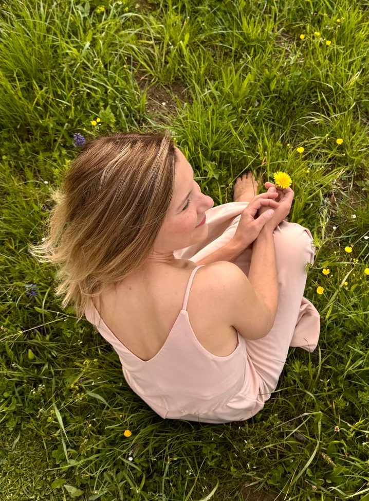 Frau in pinkem Kleid sitzt auf grünem Gras und hält eine Blume in der Hand.
