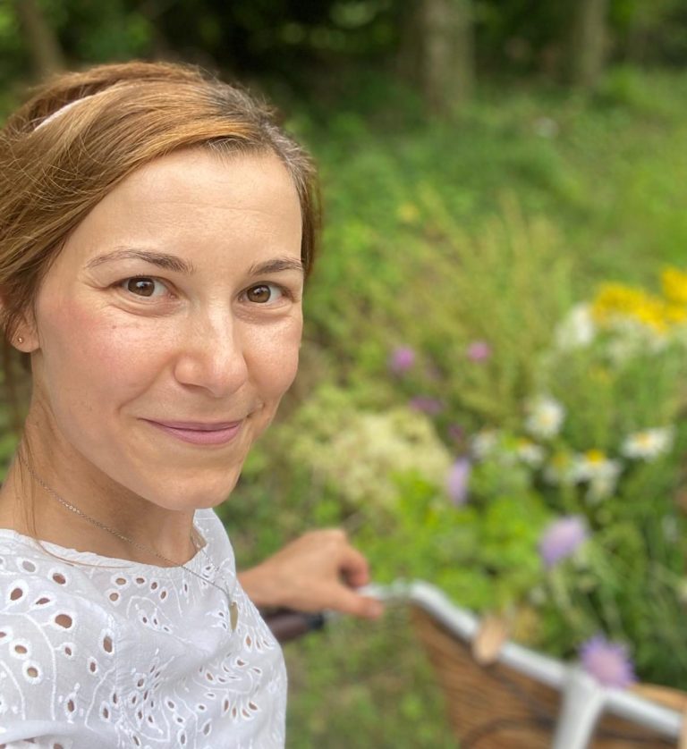 Frau mit Fahrrad in einer blühenden, grünen Umgebung lächelt in die Kamera.